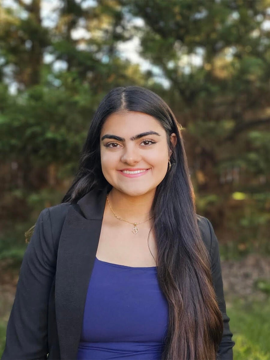 Medhnaa's headshot photo is a woman with black hair smiling with blurry green trees in the background. She is wearing a blue shirt and a black jacket