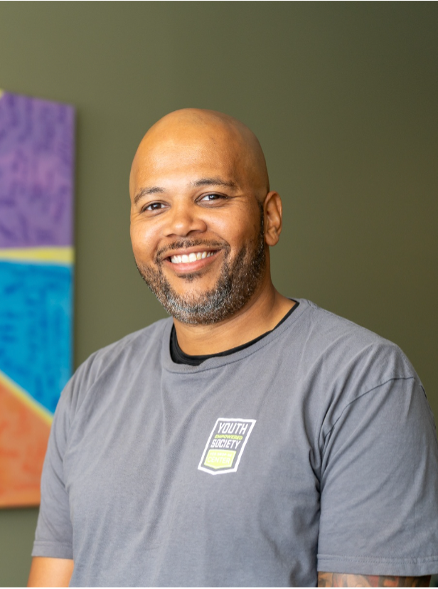 Allen Blackwell's headshot is of him in a gray t-shirt posing in front of a dark green wall.