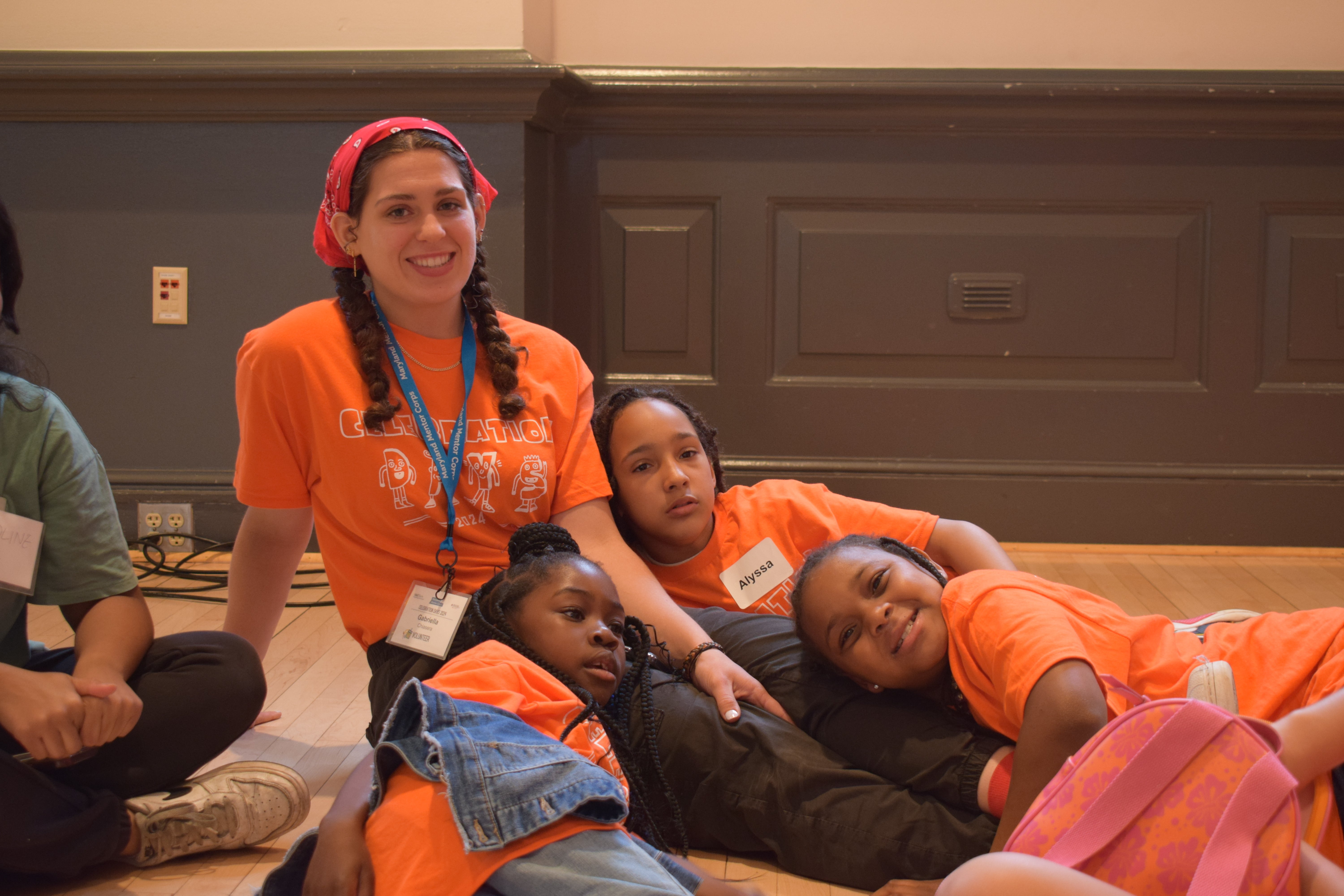 A college student is volunteering, wearing a bright orange tshirt. She is smiling and has three kids all sitting right at her feet, also wearing orange tshirts. 