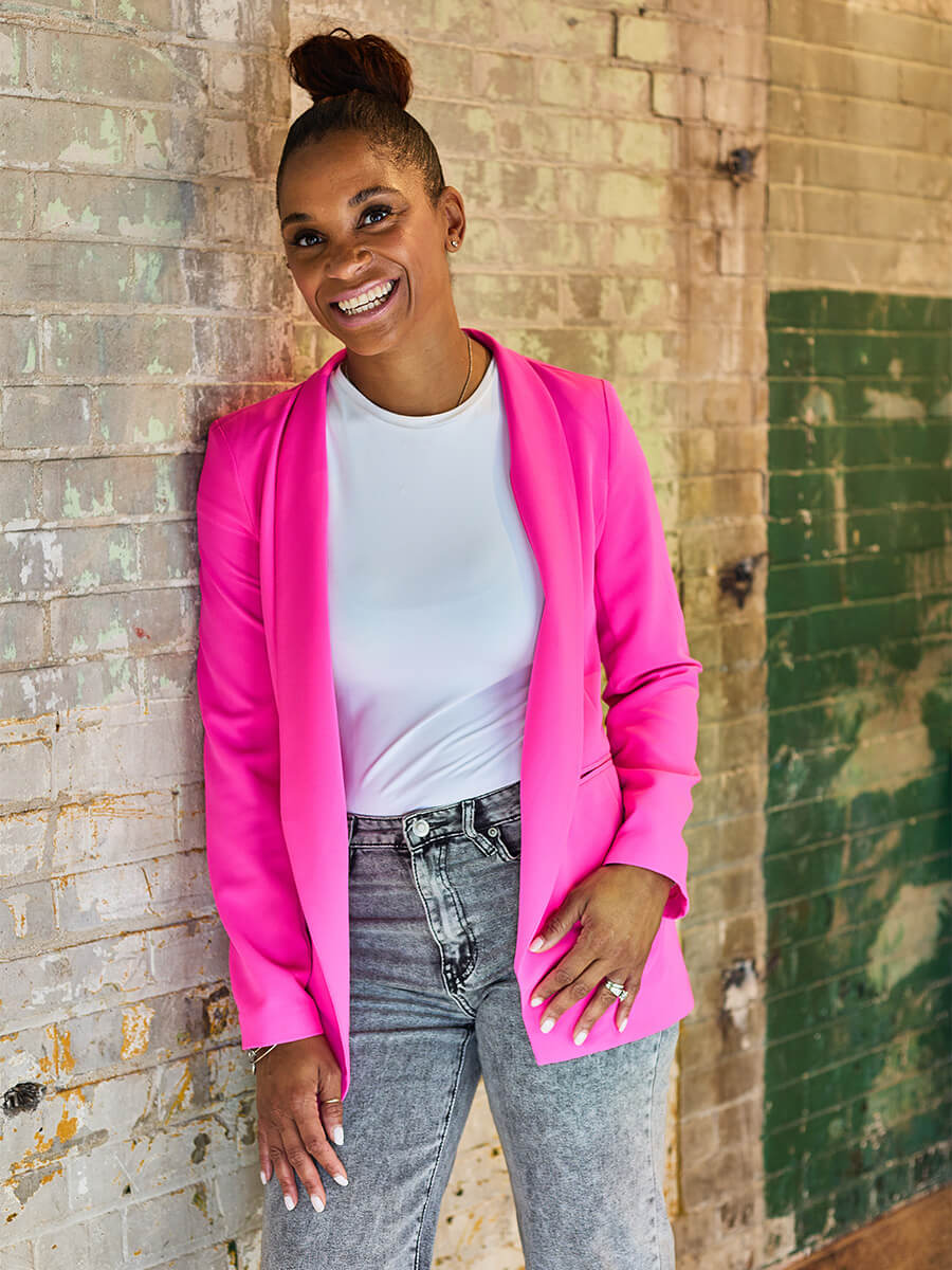 Tanasha's headshot features her smiling against a brick wall wearing a white shirt and hot pink blazer