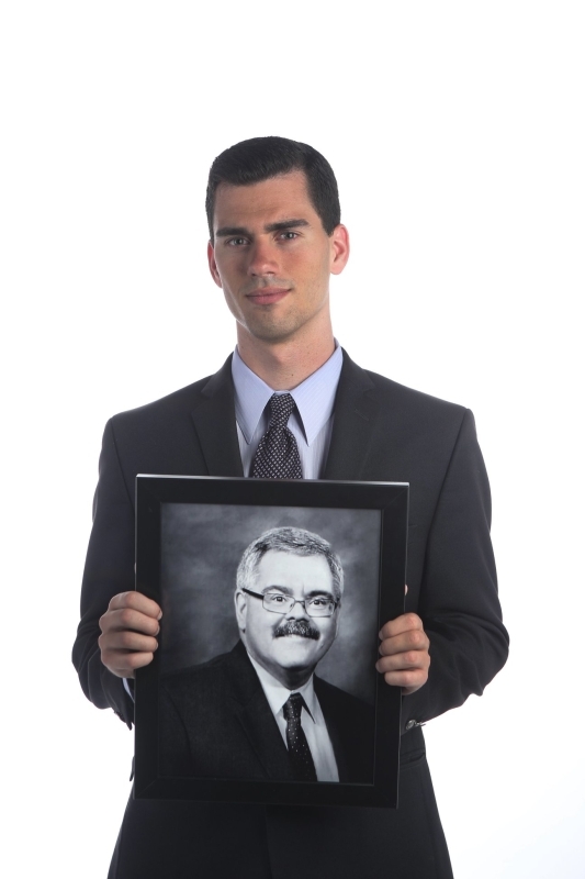photo of Matthew standing in a suit, holding a picture frame with a photo of his Father