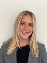 Emma Rubino is wearing a grey blazer and black shirt, and is smiling at the camera. 