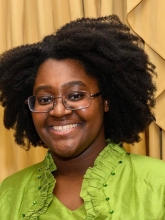Folashade headshot. She's wearing and green shirt in front of a mustard yellow background.