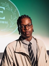 Natheer Muwonge's headshot. He's wearing a tan jacket and black tie with a green background. 