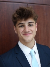 Blake Simonds headshot features him in a blue suit with a light blue tight posed with a brown background. 