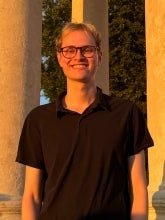 Headshot of Jakob Sandberg. He has blonde hair and a black collared shirt with black framed glasses. There are three pillars behind his head with a sunset colored overcast. 