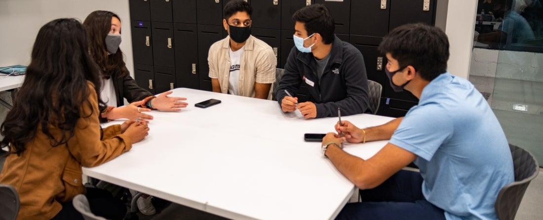 A group of students are all sitting around a table and discussing. They are all wearing masks. 