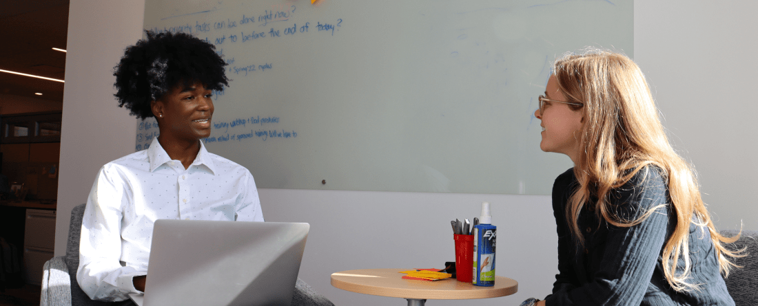 Two students sitting in chairs discussing a topic with a whiteboard behind them