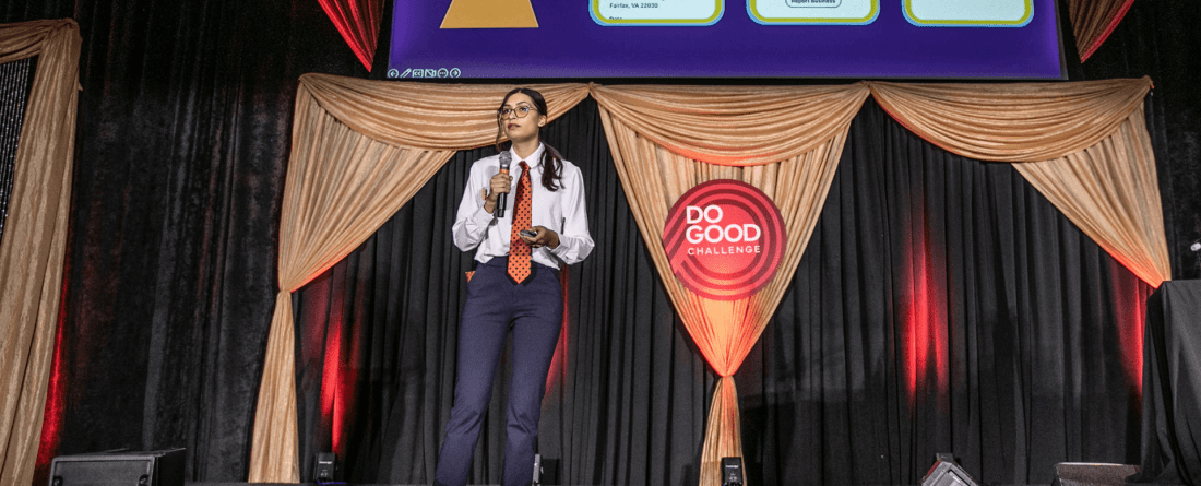 Kat Close holding a mic on stage at the Do Good Challenge with gold drapes in the background 