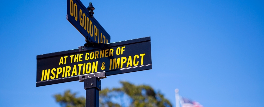 A cross street sign with the words, "At the corner of inspiration and impact on it." A bright blue sky is behind the sign. 