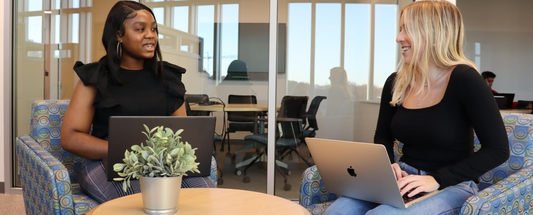 Two students are facing each other in office chairs with computers on their laps. 