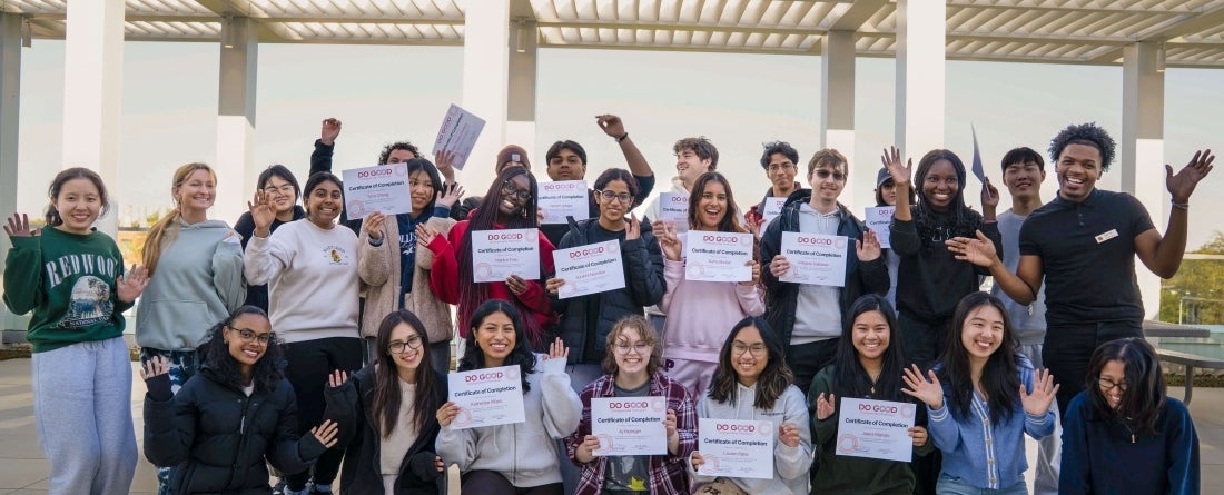 A group of students pose in three rows with their hands up and certificates in their hands during a sunny day on a rooftop.