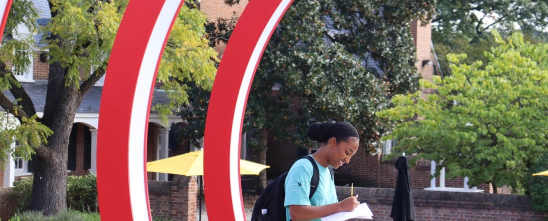 A student with a light blue shirt stands near two large red semi circle rings while writing on a sheet of paper smiling. Green trees and a yellow umbrella color the background.