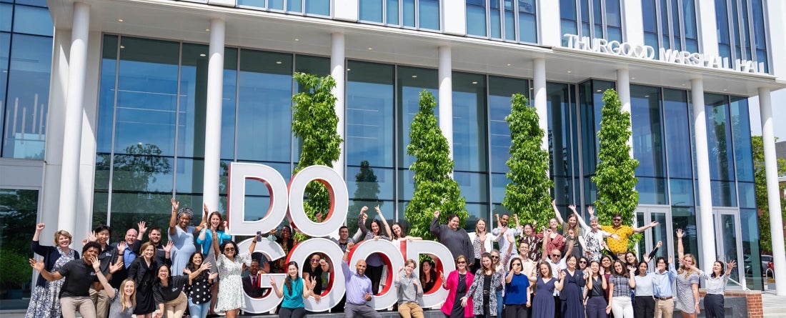 A group of faculty and staff stand in front of a large sculpture that says "Do Good" with arms raised and big smiles on a nice sunny day. 