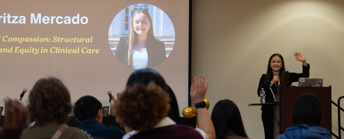 A student presents at a podium, engaging an attentive audience as several participants raise their hands to contribute to the discussion.