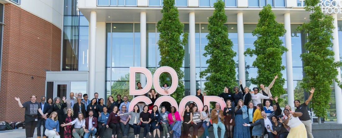 A group of people pose with smiles and hands raised in front of sculpture that says "Do Good"